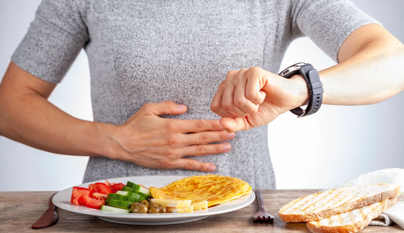 Concept of intermittent fasting with a woman sitting hungrily in front of the food and watching the clock to make sure she breaks the fast at the right time.
