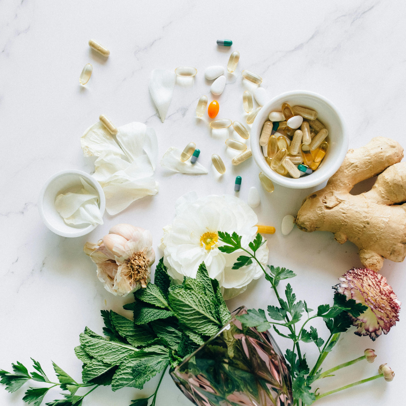 A marble surface displays vibrant flowers, fresh herbs, and colorful organic vitamin supplements in pill form