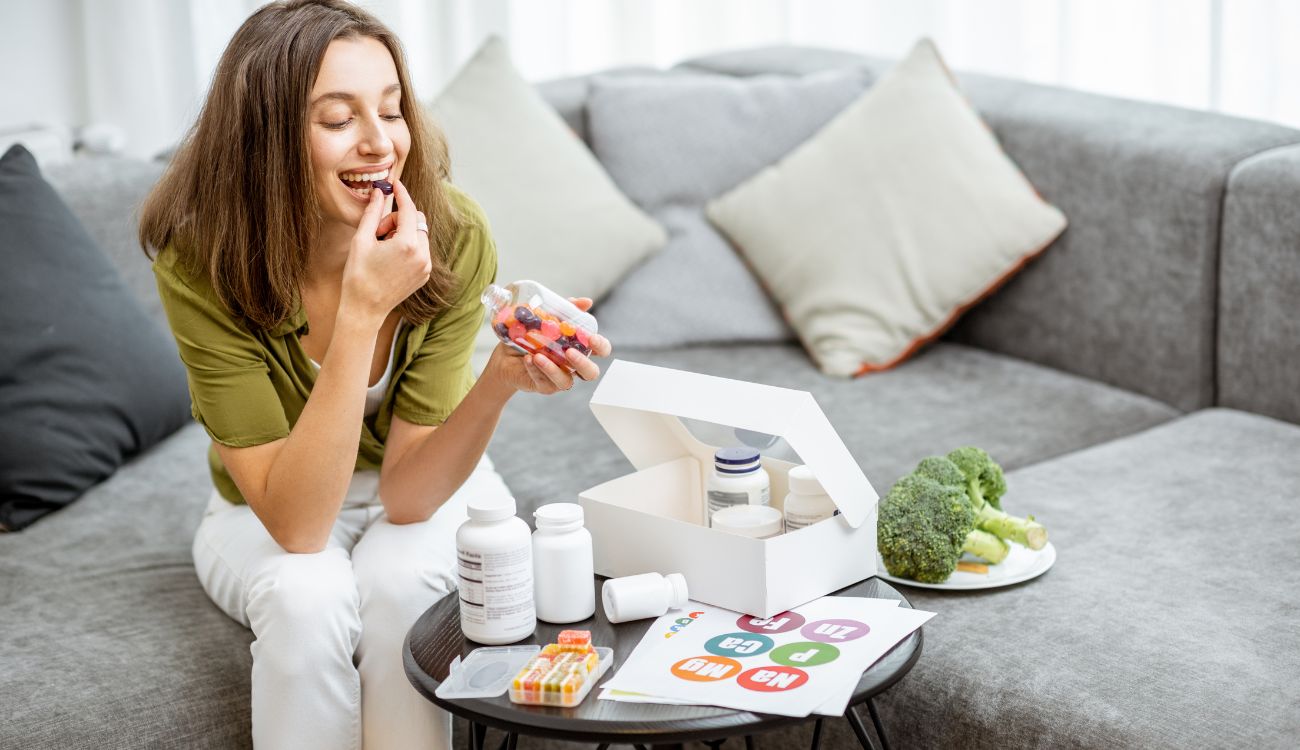 A woman sitting on a couch, taking vitamins from a box alongside other wellness items.